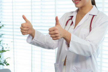 Young Caucasian female pediatrician wearing white lab coat with stethoscope giving thumbs up expressing positivity. Professional medical expert demonstrating confidence in healthcare services.