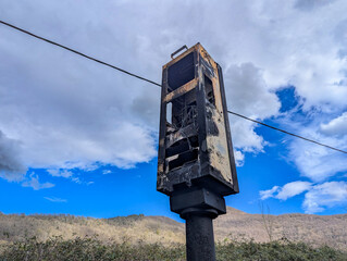 Radar de velocidade carbonizado à beira de uma estrada em contraste com a serenidade do céu parcialmente nublado