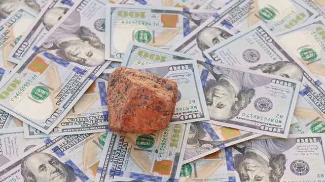 A rough orange granite rock spins on a table covered with 100-dollar US bills, completing a full rotation, highlighting the connection between natural minerals and financial wealth.