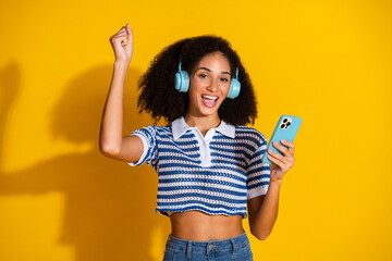 Young woman listening to music on headphones while holding a smartphone and smiling joyfully against a vibrant yellow background