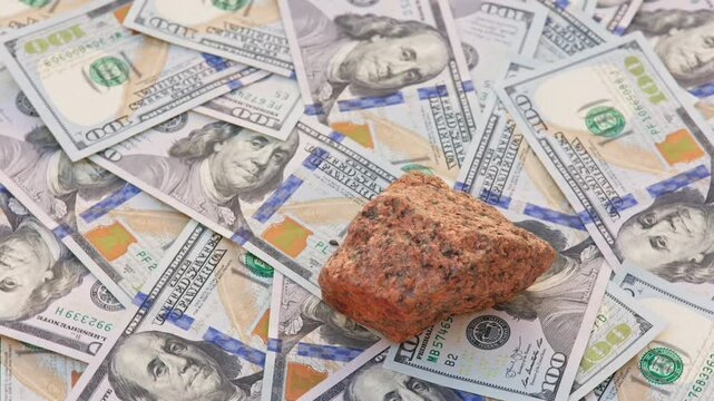 A rough orange granite rock spins on a table covered with 100-dollar US bills, completing a full rotation, highlighting the connection between natural minerals and financial wealth.