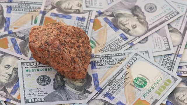 A rough orange granite rock spins on a table covered with 100-dollar US bills, completing a full rotation, highlighting the connection between natural minerals and financial wealth.
