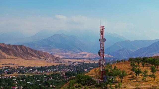 Drone footage showing a mountain village near with a telecom tower on a dry hill. The drone slowly passing by a tower with distant mountains in the background at late summer day.
