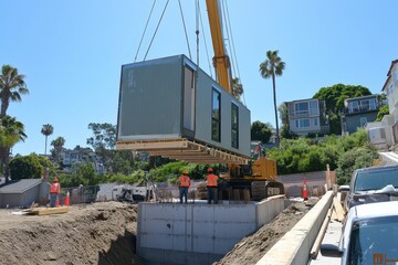 Construction workers operate a crane to place a modular home on a foundation in a sunny area