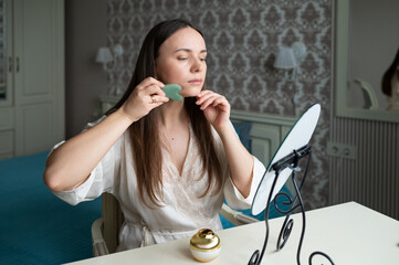 A young woman in a silk robe uses a jade gua sha facial tool while sitting at a vanity mirror in a...
