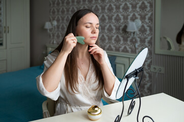 A young woman in a silk robe uses a jade gua sha facial tool while sitting at a vanity mirror in a...