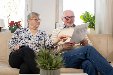 couple of elderly migrants sit on sofa with papers in their hands. They read letters from...