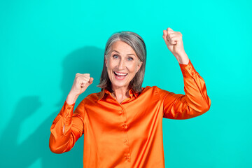 Excited elder woman in elegant orange blouse celebrating against vibrant turquoise background