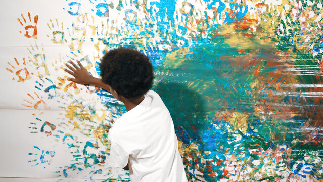 Back view of happy african boy painted the stained wall with colorful hand while wearing casual white shirt in art lesson.Smart student use hand print to make creative artwork. Education. Edification.