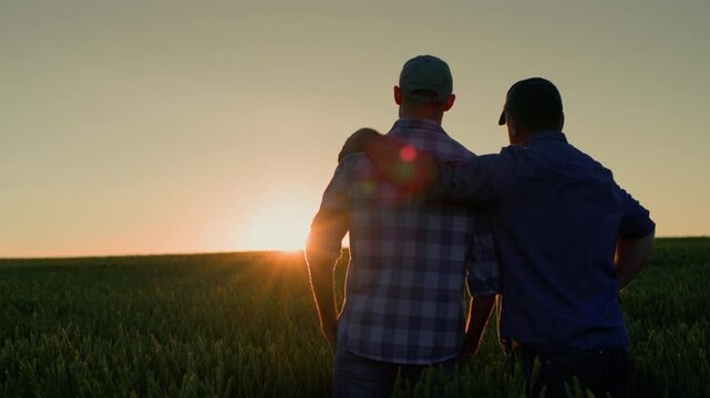 Father and son in a field at sunset. The son is slightly in front, wearing a brightly colored plaid shirt and baseball cap. His arm is around the shoulders of the man behind him.