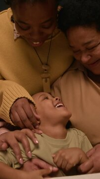 Vertical shot of happy Black family playing with cheerful little boy touching kids nose during holiday celebration at home, handheld camera
