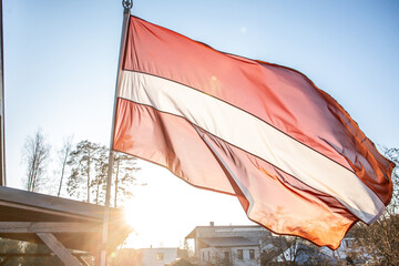 The Latvian flag waves prominently with red and white stripes, set against bare trees, rooftops,...