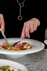 A close-up of a person using a fork and knife to eat a fresh salad with fried chicken pieces, cherry tomatoes, and grated cheese on a white plate.