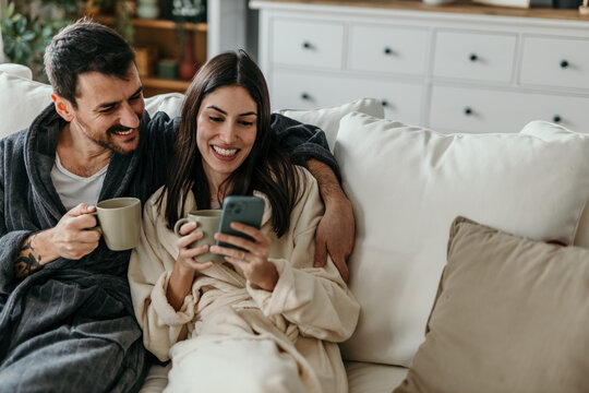 Couple relaxing on sofa, drinking coffee and using smartphone