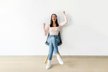 Overjoyed young woman screaming raising two arms fists up and looking at camera, sitting in chair over white wall background, full length
