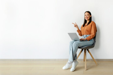 Happy woman working online on laptop and pointing aside at free space, sitting in chair near white...