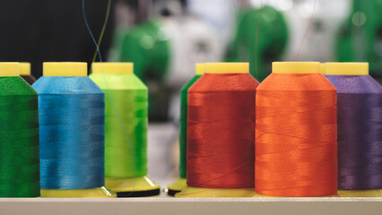 A row of colorful spools of thread are displayed on a shelf. The spools are of various colors, including green, yellow, orange, and purple. The spools are arranged in a neat row
