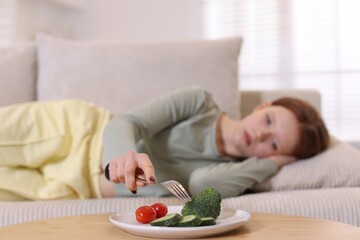 Sad teenage girl lying on sofa near table with vegetables at home, selective focus. Eating disorder