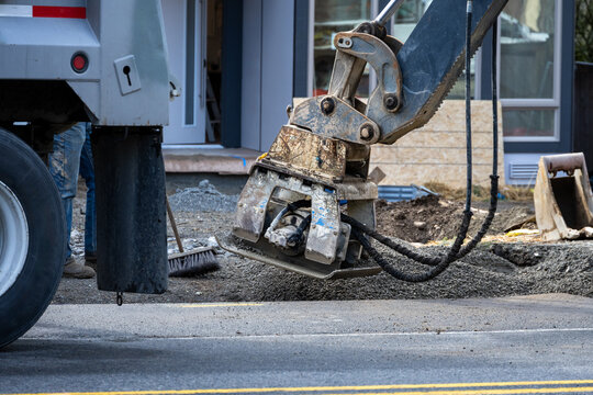 Roadworks covering trench dug for underground utilities with gravel and compacting with vibrating tamper attachment on excavator machine, new housing development construction project
