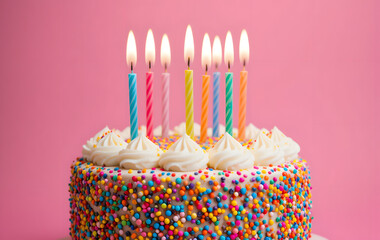 Colorful birthday cake decorated with sprinkles, whipped frosting, and lit candles on a pink background, creating a cheerful and festive celebration scene