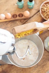 A person adds freshly grated lemon zest into a mixing bowl in a cozy kitchen. Ingredients like eggs and nuts are visible, indicating dessert preparation