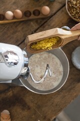 A hand pours bright yellow zest into a mixing bowl while using a stand mixer in a cozy kitchen. The workspace features wooden surfaces and baking ingredients