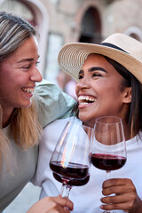 Vertical. Two cheerful multi-ethnic female friends are happily toasting with glasses of red wine while enjoying their vacation together in an outdoor setting in an italian city
