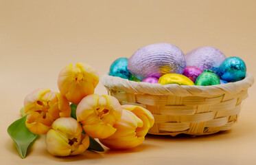 An Easter composition featuring a wicker basket filled with colourful foil-wrapped candy eggs, with soft pastel beige-yellow tulips, set against a minimalistic pastel beige-yellow background. 