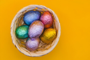 A festive Easter composition featuring a wicker basket filled with colourful foil-wrapped candy eggs, set against a vibrant yellow background. For cards, wallpapers, Easter designs.