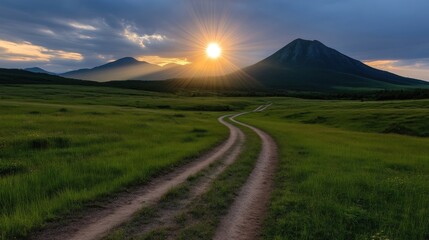 Naklejka premium Scenic dirt road winding through a lush meadow, leading to majestic mountains at sunset. Sunlight beams through clouds