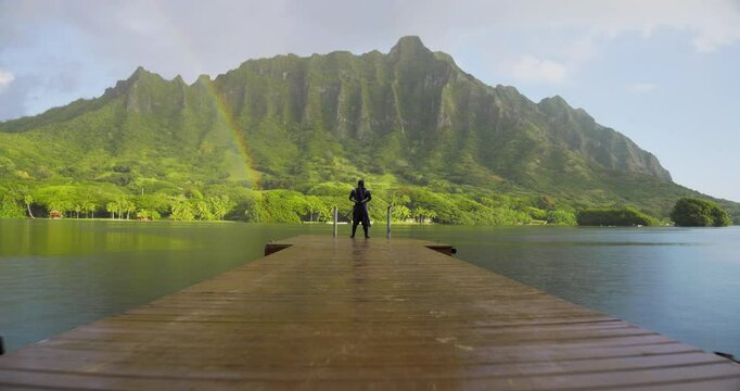 Cinematic push in on lone ninja knight standing on dock of tropical paradise with rainbow, mask falling and man laughing