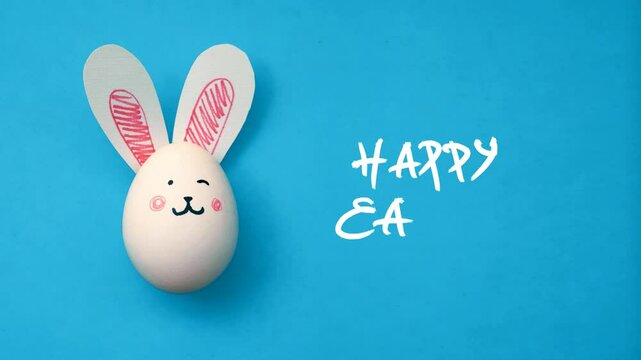 Horizontal video. Chicken white egg with cute bunny muzzle, paper rabbit ears and hand drawn text Happy Easter on light blue. Frame by frame, stop motion animation. Concept of egg hunt, spring, gift