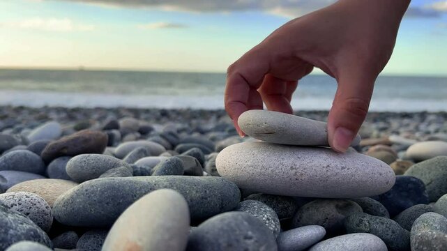 Horizontal 4K video close up. Female hand builds small rock tower on pebble beach. Blue sky. Light stones. Sun, sea waves, coast line. Concept of rest, ocean shore zen, calm, yoga, travel. Sunny day