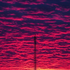 Obraz premium Crimson and magenta sunset sky with layered clouds, a tall communication tower silhouetted against the vibrant colors.