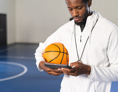 Coach holding basketball and using tablet on indoor court, planning strategy
