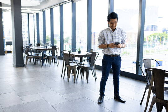 Businessman in office cafeteria using smartphone, standing near large windows, copy space