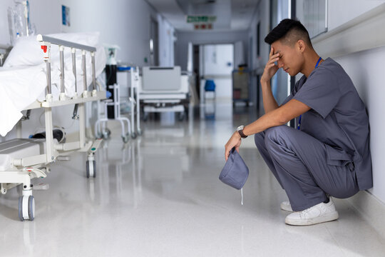 Exhausted healthcare worker in scrubs sitting in hospital corridor, holding cap, copy space