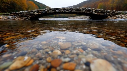 Obraz premium Clear river flowing under ancient stone arch bridge. Autumn foliage beyond