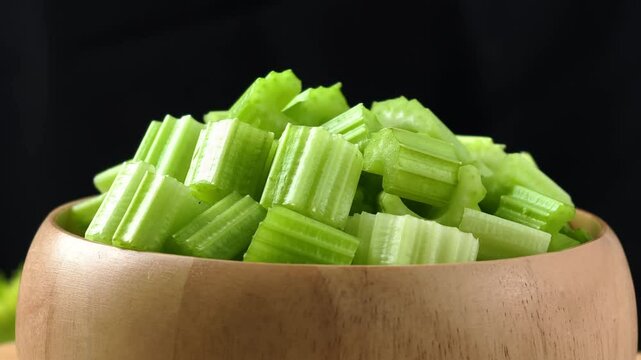 Fresh chopped celery in a wooden bowl with whole celery stalks on a rustic dark surface