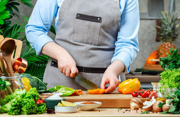 Chef cuts orange paprika into slice. Cozy kitchen with wooden table, kitchenware, vegetables, herbs and ingredients for cooking. Healthy vegan eating, culinary, recipes, food blogging