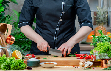 Chef in uniform cuts hot green chili pepper. Cozy kitchen with wooden table, kitchenware, vegetables, herbs and ingredients for cooking. Healthy vegan eating, culinary, recipes, food blogging