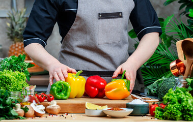 Chef holding green, red, yellow paprika in hands. Cozy kitchen with wooden table, kitchenware, vegetables, herbs and ingredients for cooking. Healthy vegan eating, culinary, recipes, food blogging