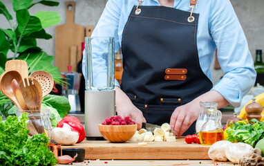 Chef cuts bananas for making smoothie with raspberry and alternative milk near blender. Cozy kitchen with wooden table, kitchenware, vegetables, herbs and ingredients for cooking. Healthy vegan eating