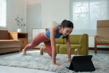 Woman in activewear performing a plank exercise while following a workout video on a tablet in a cozy living room setting. Bright natural light enhances the modern decor