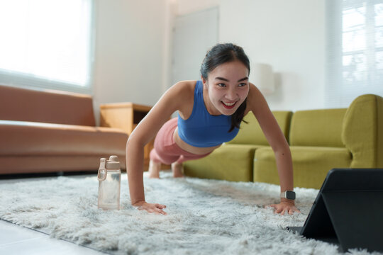 Happy sportswoman is training her upper body strength by doing push-ups on a carpet in her living room, following instructions from a fitness video tutorial on a tablet