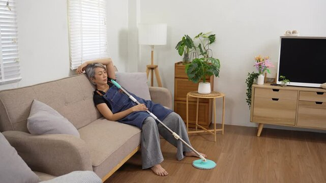 Tired senior housekeeper resting on sofa after cleaning, elderly woman exhausted from housework in cozy home