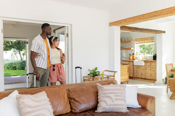 Diverse couple entering vacation home with luggage, smiling and exploring cozy living room, copy spa