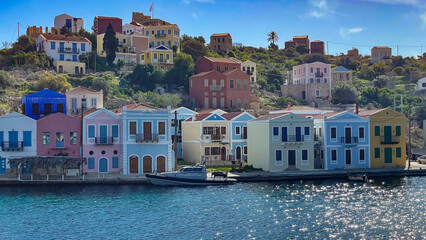 Colorful Traditional Houses On The Waterfront Of Kastellorizo Island, Greece, Built On A Hillside Overlooking The Sea, Greek Flag In The Background, Peaceful Atmosphere, Ideal For Travel And Tourism.