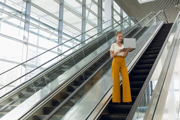 Using laptop on escalator, businesswoman multitasking in modern conference center, copy space