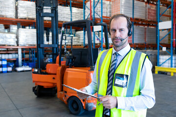 Wearing safety vest and headset, warehouse manager holding clipboard overseeing operations, copy spa